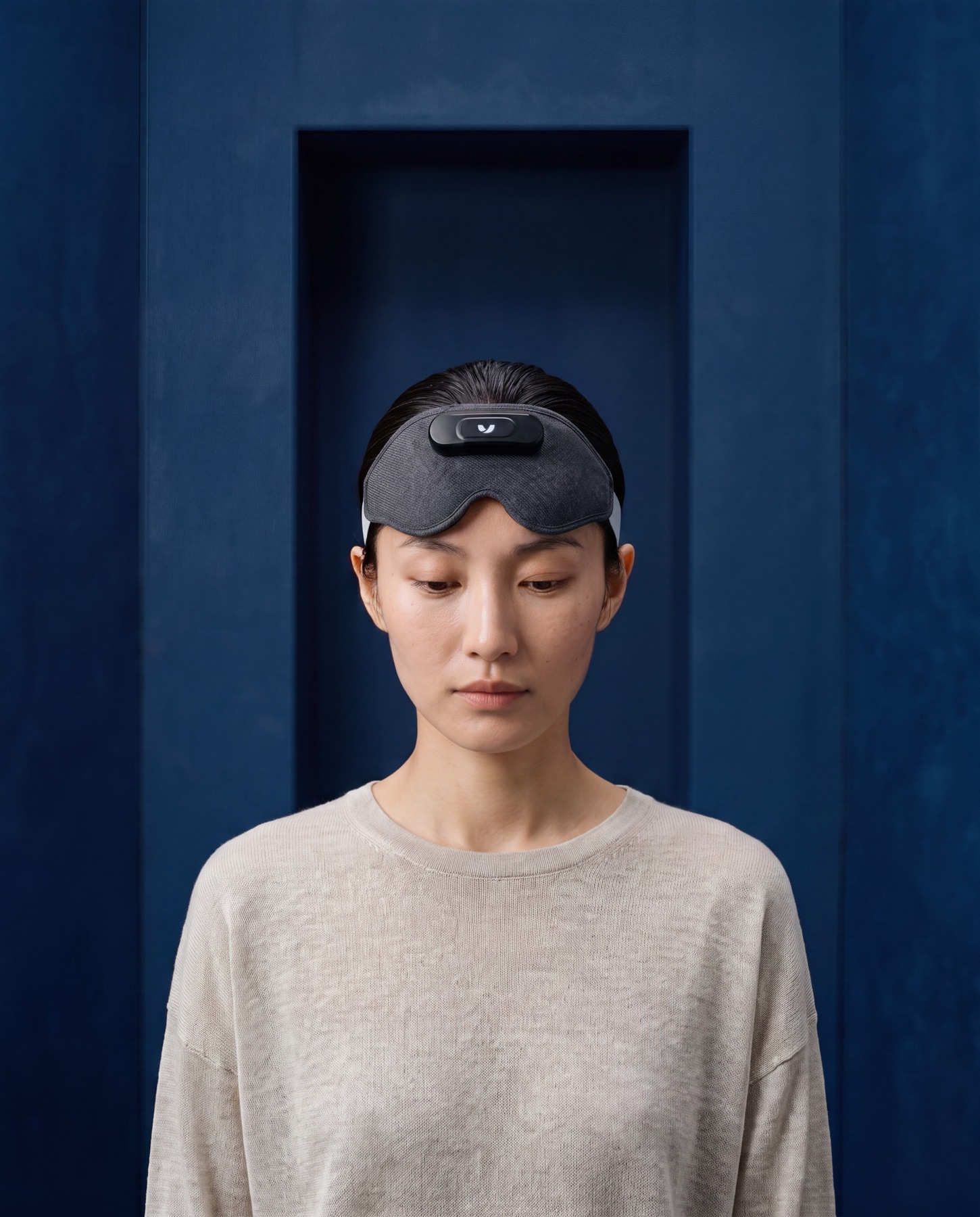 Woman with slicked-back hair and the Mask pushed up on her forehead, eyes lowered, in a navy monolithic chamber