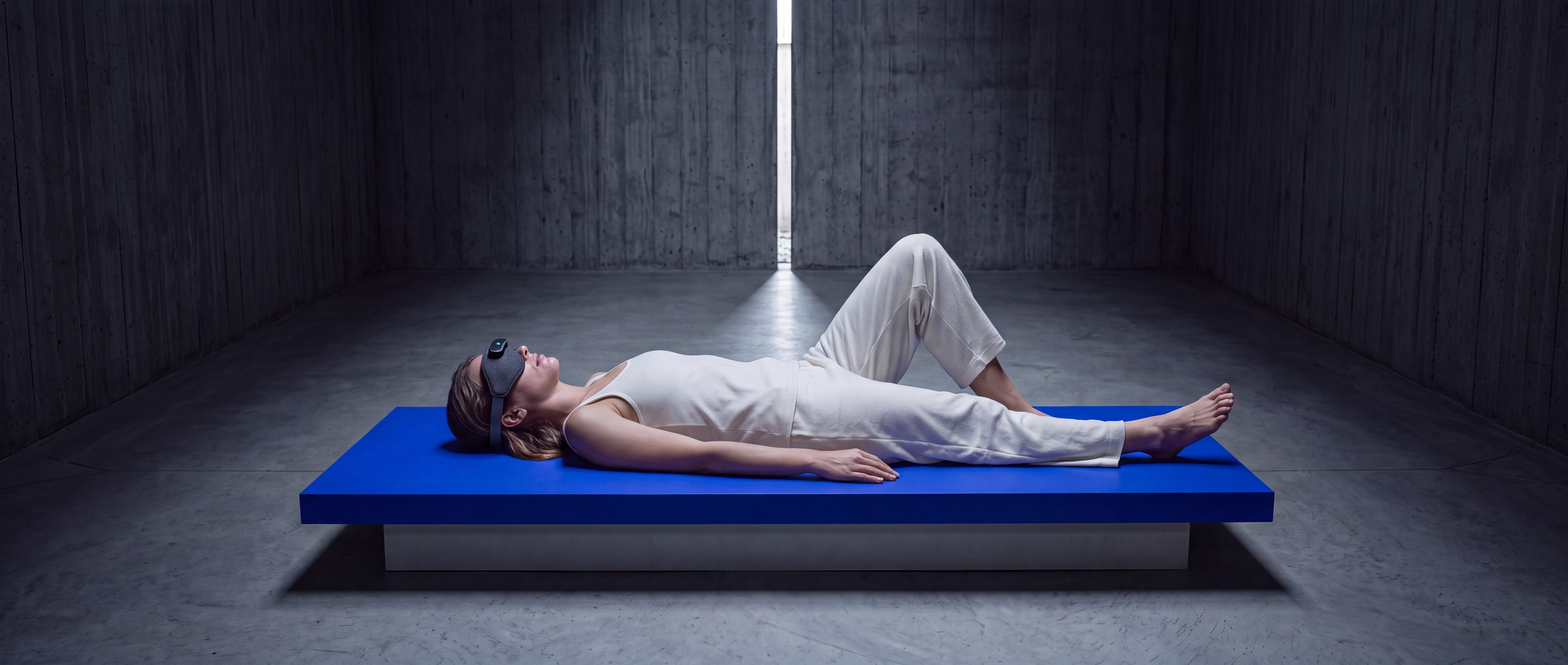 A person lying perfectly still on a floating electric-blue slab in a vast dark concrete hall, wearing the Mask over their eyes under a single shaft of light