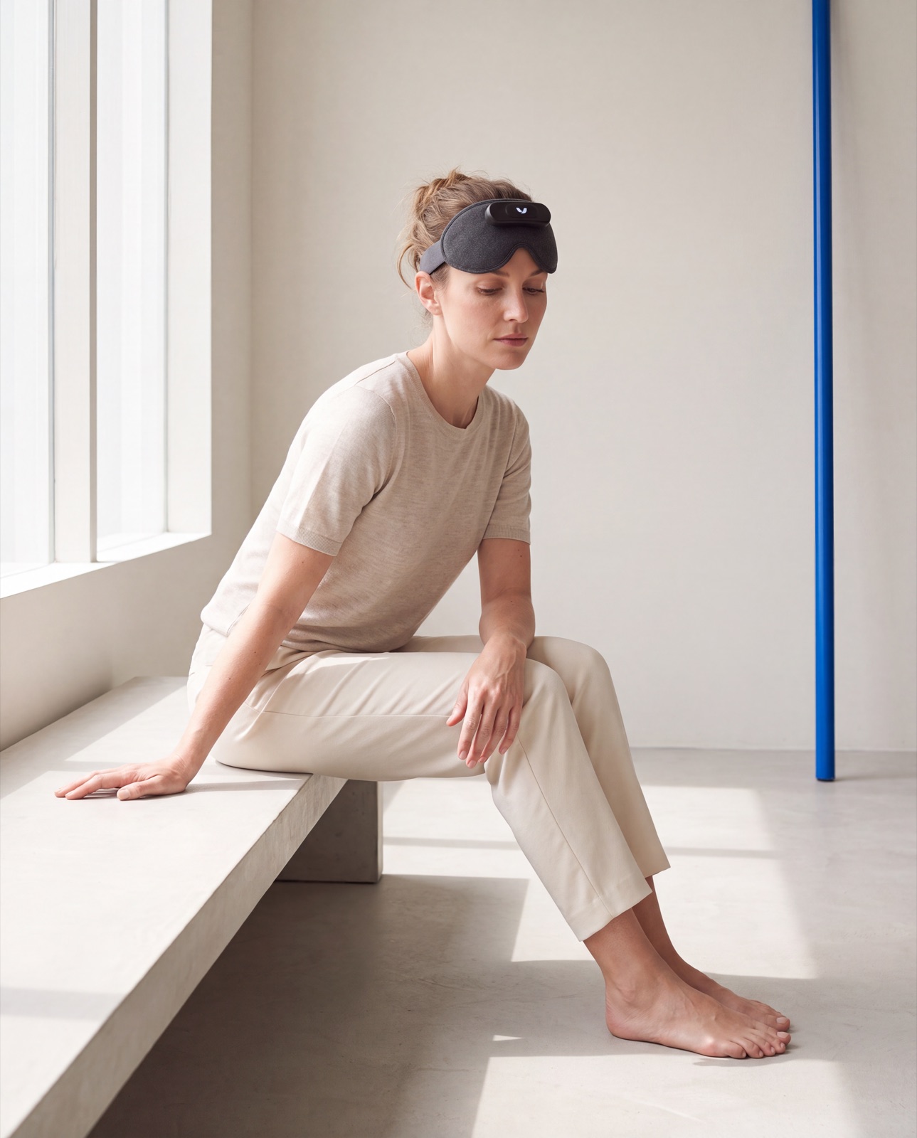 A composed woman in a cream tailored suit, the Mask pushed up on her forehead, standing in a concrete gallery beside a cobalt-blue panel